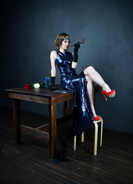piękna kobieta w niebieskiej sukni wieczorowej w czarnych rękawiczkach z papierosem i butelką siedzącą samotnie na stole - Zdjęcie, obraz