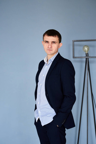 Young brunette man, wearing dark blue suit and light blue shirt, standing in front of blue wall with cinema lamp, hands in the pockets. Businessman, posing for resume picture. Corporate culture - Фото, изображение