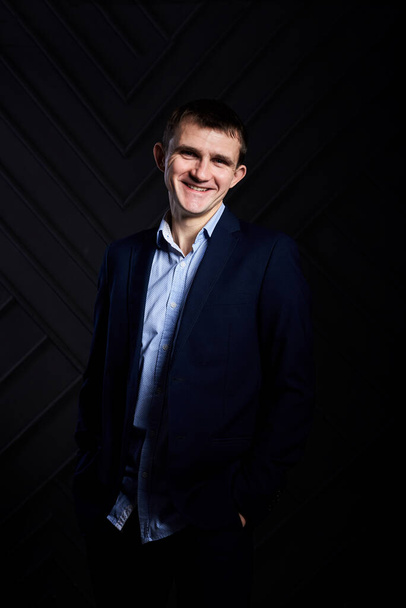 Young brunette man, wearing dark blue suit and light blue shirt, posing for picture in front of grey black wall in photo studio, smiling, laughing. Businessman Corporate culture. Happy Emotions. - Fotó, kép