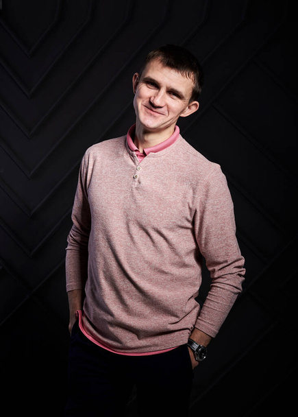 Young brunette man, wearing casual light pink jumper, posing for picture in front of black wall in photo studio, smiling, laughing, hands in pockets. Businessman Corporate culture. Happy Emotions. - Фото, изображение
