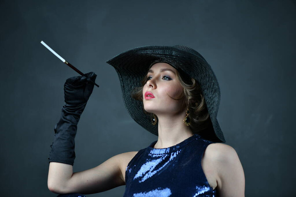 piękna kobieta w niebieskiej sukni wieczorowej z czarnymi rękawiczkami i czarnym kapeluszem i fajką - Zdjęcie, obraz