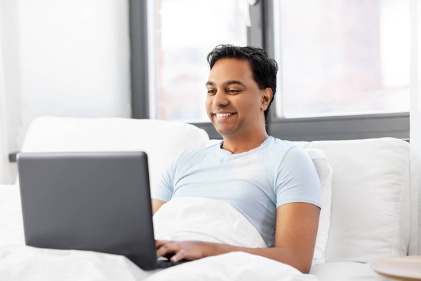 happy indian man with laptop in bed at home - Photo, image