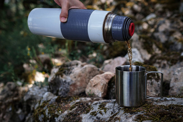 キャンプ用マグカップでは、女性の手が自然の魔法瓶からお茶を注ぎます。休憩、休息、旅行のアイデアとコンセプト - 写真・画像