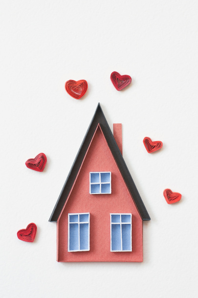 Rood papieren huis met blauwe ramen met rode harten. Klein huis gemaakt in quilling techniek op witte achtergrond. Handgemaakt van papier. Blijf thuis. Huis zoete huis.Concept van de liefde in huis - Foto, afbeelding
