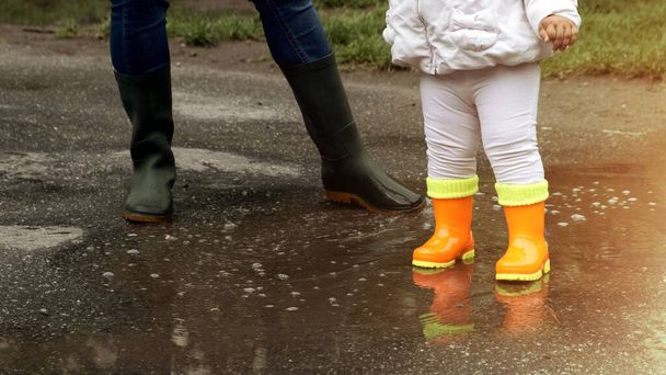 Kinderorangefarbene und erwachsene grüne Gummistiefel in einer Pfütze nach Regen an einem sonnigen Tag. - Foto, Bild