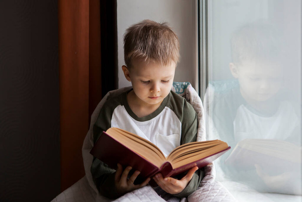 Χαριτωμένο μικρό αγόρι διαβάζει το βιβλίο, ενώ κάθεται στο περβάζι παράθυρο. - Φωτογραφία, εικόνα