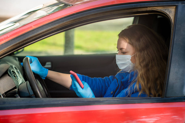 A woman driving a car in protective medical mask and gloves is talking on phone. Lifestyle and safe drive during a pandemic coronavirus. Protect driver and passengers. New normal and modern reality - Foto, Bild