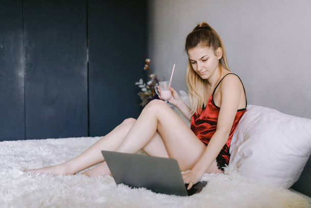 Νεαρή γυναίκα με milkshake χρησιμοποιώντας φορητό υπολογιστή στο κρεβάτι - Φωτογραφία, εικόνα