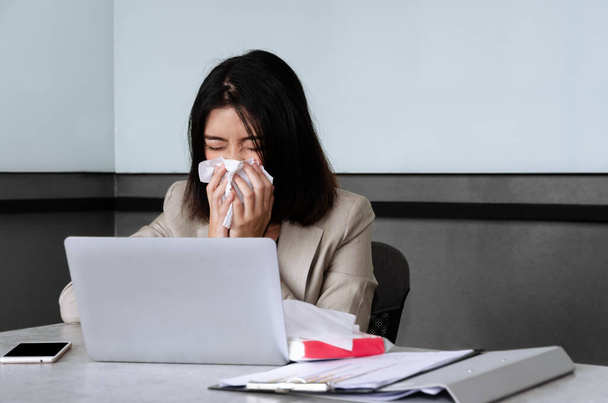 Hasta iş kadını, ofis masasında dizüstü bilgisayarla oturmuş toplantı odasında çalışırken hapşırıyor ve yüz dokusuna öksürüyor. İş yerinde Coronavirus covid-19 yayılması ve riski - Fotoğraf, Görsel