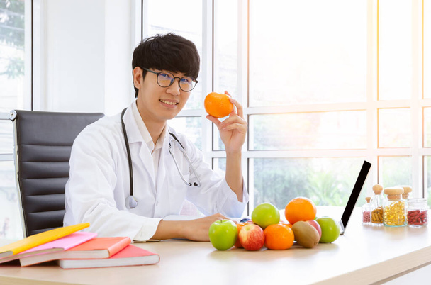 Férfi táplálkozási szakértő fehér köpenyben, narancssárga gyümölcsöt tart a klinikán, friss gyümölcsökkel és vitaminokkal az asztalán. Egészséges táplálkozás gyümölcs és C-vitamin koncepció az influenza megelőzésére. - Fotó, kép