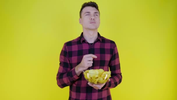 男はガラスボウルからポテトチップスを食べ、黄色の背景で味を楽しんでいます - 映像、動画
