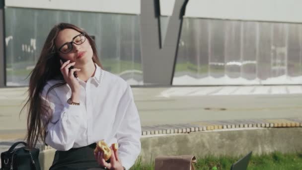 Kaunis Business nainen syö croissant puhuessaan matkapuhelimeen ja hymyillen ulkona - Materiaali, video