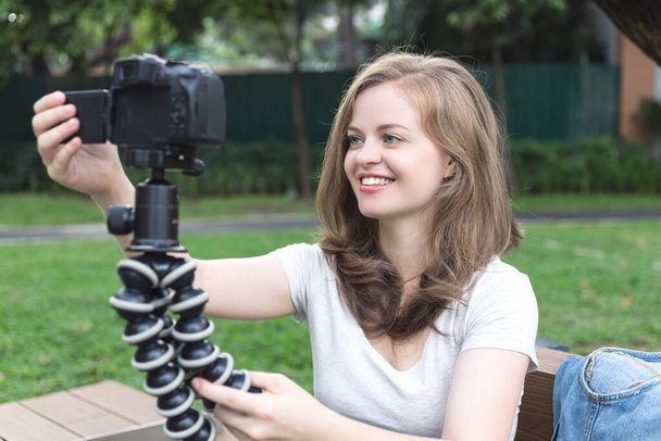 Sonriente joven caucásica chica mujer haciendo un video blog (vlog) con cámara afuera en verde parque
 - Foto, Imagen