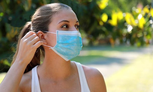 Girl in the park puts on a medical mask. Young sporty woman wearing surgical mask in city park. Copy space. - Photo, Image