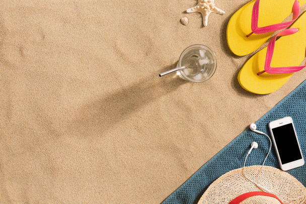 Summer vacation composition. Flip flops, hat, smartphone and glass of water on sand background. Travel vacation concept. Summer background. Border composition made of towel - 写真・画像