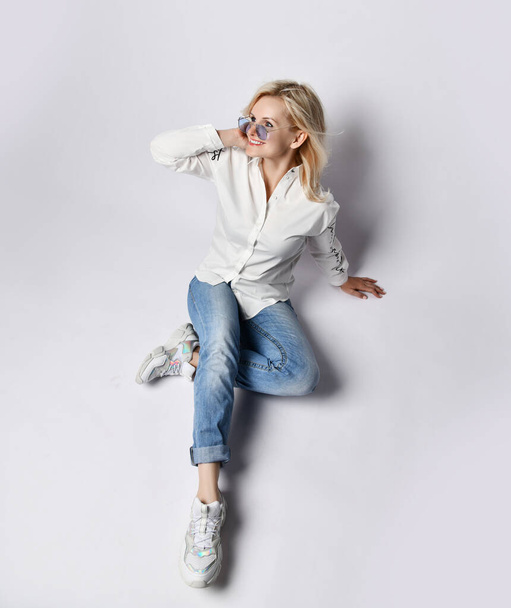 Top view of young joyful blond woman in casual clothing, white sneakers and sunglasses sitting on floor and smiling - Φωτογραφία, εικόνα