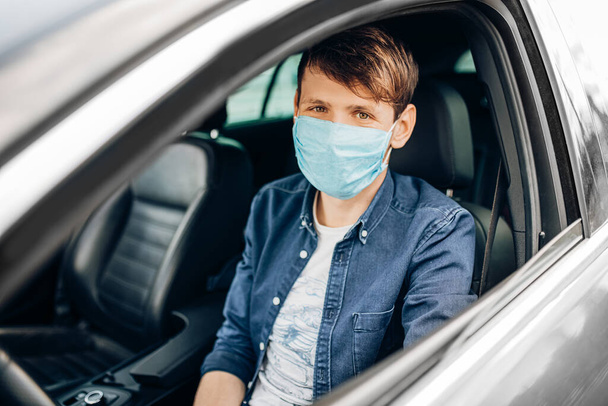 A young man in a protective medical mask, sitting at the wheel of a car, the driver is driving. Quarantine, coronavirus - Φωτογραφία, εικόνα