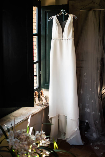 Egy gyönyörű fehér esküvői ruha. Esküvői ruha lóg az ablak mellett. jó fény. - Fotó, kép