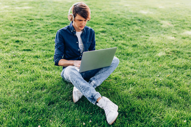 A young handsome man is sitting outside on the grass with a laptop and listening to music with headphones - Фото, зображення