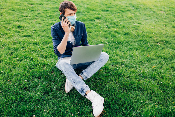 A young man in a protective medical mask on his face is sitting on the grass in a Park with a laptop and talking on a mobile phone - Photo, Image