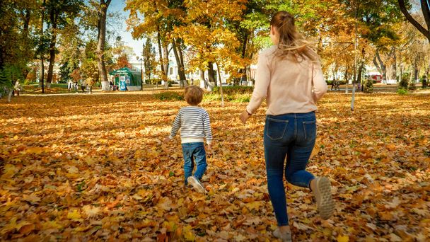 Šťastná mladá matka se svým malým synem běží na padlých žlutých listech v podzimním parku - Fotografie, Obrázek
