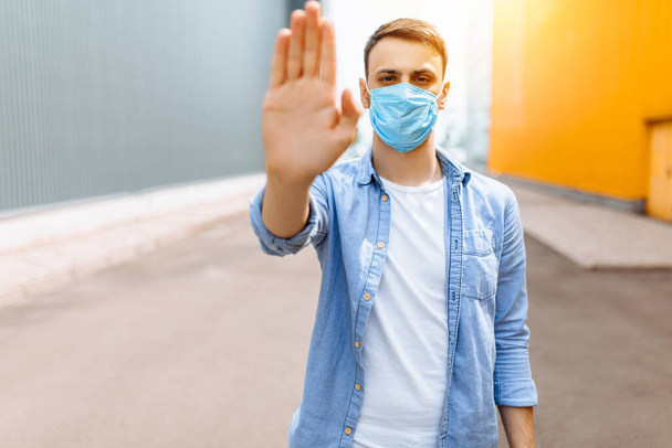 A young man in a protective medical mask makes a stopping hand gesture while standing in the city. Quarantine, epidemic, coronavirus - Фото, зображення