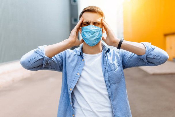 A confused panicked man in a medical protective mask, holding his head in fear, standing in the open air in the city. Coronavirus, an epidemic - Foto, imagen