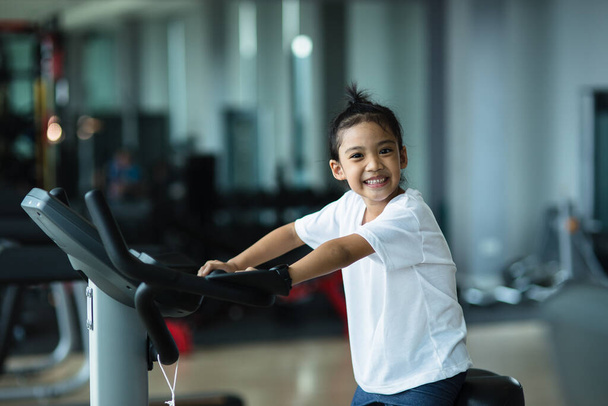 Ein kleines Mädchen mit Heimtrainer in der Turnhalle. Fitness-Frau nutzt Airbike im Fitnessstudio. - Foto, Bild