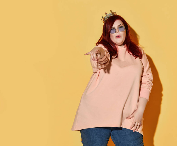 Flirting red haired overweight woman in casual clothing and golden crown on head points finger at us - Foto, afbeelding