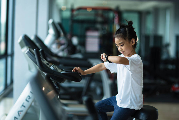 Mała dziewczynka patrzy na inteligentny zegarek po treningu na siłowni. Zdrowa koncepcja. Kobieta po sesji treningowej sprawdza wyniki na zegarku.Koncepcja sportowa dla dzieci - Zdjęcie, obraz