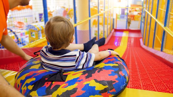 Netter kleiner Junge reitet auf aufblasbarem Donut-Ring in Kinderspielzimmer im Einkaufszentrum die Rutsche hinunter - Foto, Bild
