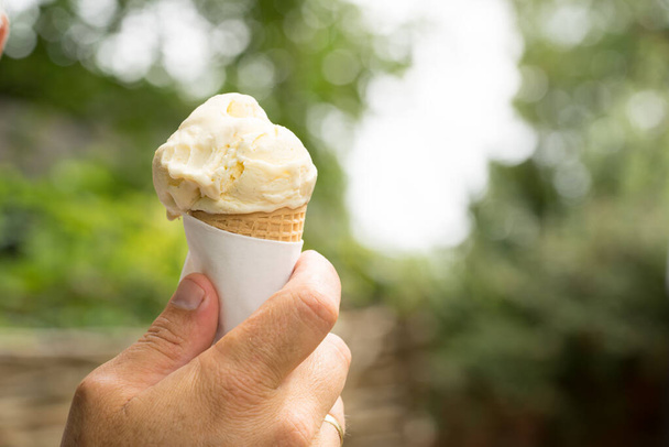 Um cone com uma colher de sorvete de baunilha realizada em uma mão masculina causasiática ao ar livre com um jardim no fundo - Foto, Imagem