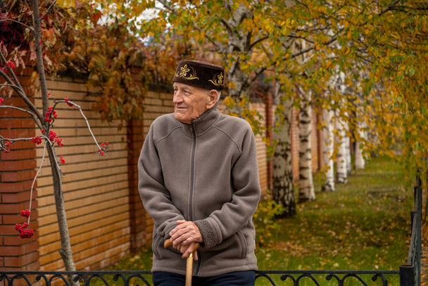 Half length portrait of an elderly Asian man Muslim in skullcap sitting with a stick on the fence by the house - Фото, зображення