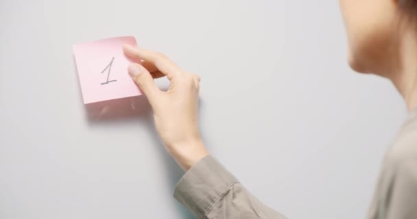 Young woman sticking pink reminder stickers with numbers 1 2 3 on a gray board. Copy space. - Video