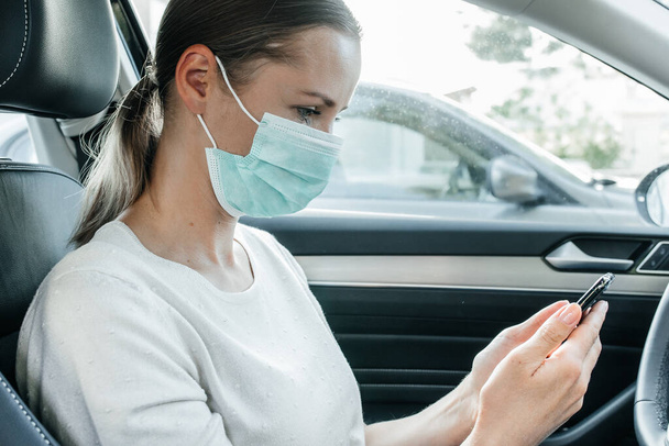 Coronavirus önleme. Koruyucu maske takan güzel beyaz bir kız telefonunda mesajlar okuyor ve araba kullanıyor. Covid-19 salgını. Salgın. - Fotoğraf, Görsel