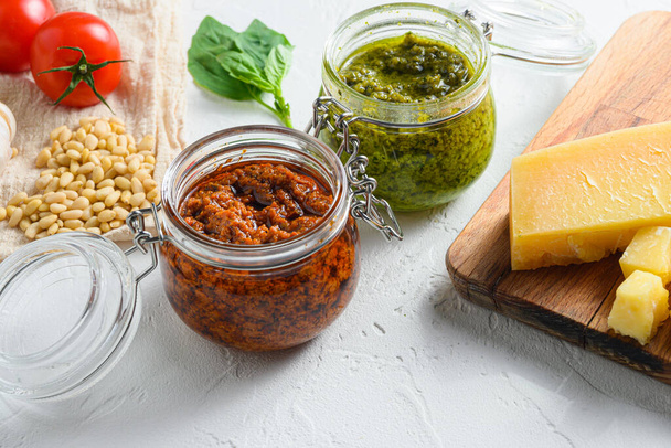 Glass jars with red and green pesto and cooking ingredients Parmesan cheese, basil leaves, pine nuts, white stone background, close up side view. - Zdjęcie, obraz