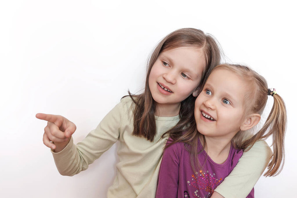 Two little girls in light and purple sweaters on a white gray background look sideways where one of them points with her finger - Zdjęcie, obraz
