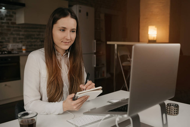 Távoli munka. Egy fehér nő fülhallgatóval a laptopján. Egy fehér inges lány jegyzetel egy online üzleti eligazításon a kényelmes otthoni munkahelyén.. - Fotó, kép