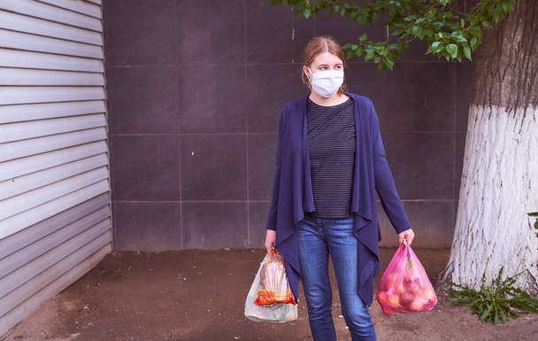 Uma jovem com uma jaqueta azul fica em um fundo escuro em uma máscara médica e mantém sacos de comida do supermercado. Foto muito alta qualidade
 - Foto, Imagem