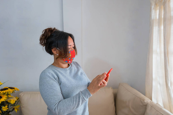 Латиноамериканка стоїть з маскою обличчя у вітальні і дивиться на мобільний телефон. - Фото, зображення