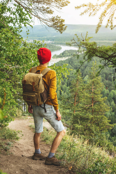 Podróżnik turystyczny z plecakiem i czerwonym kapeluszem stoi na krawędzi klifu i spogląda na zieloną dolinę z rzeką. Koncepcja wolności i jedności z naturą - Zdjęcie, obraz