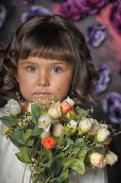 little brunette girl with curls with a white sweater with flowers in her hands on a floral background in the studio - Foto, immagini