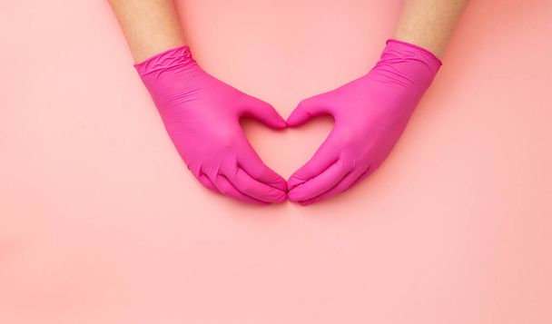 Top view, female hands in protective gloves folded together in form of a heart on pink background. - Zdjęcie, obraz