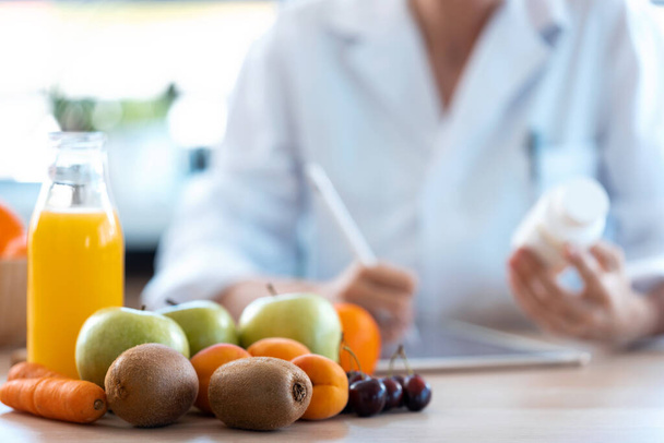 Close-up of woman nutritionist doctor writes the medical prescription for a correct diet on a desk with fruits, pills and supplements. - Zdjęcie, obraz