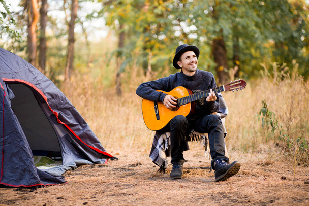Jeune homme souriant en chapeau noir assis près d'une tente touristique et jouant de la guitare dans la forêt d'automne - Photo, image