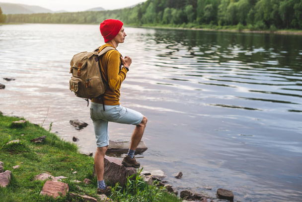 A tourist traveler with a backpack and a red hat are standing by the river with a beautiful reflection from the water in the forest at sunset. Concept active recreation and unity with nature - 写真・画像