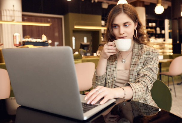 Portrait of confident young businesswoman in smart casual wear drinks coffee and working on laptop in a cafe interior - Photo, Image