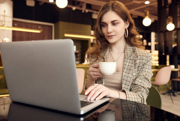 Retrato de la joven empresaria confiada en ropa casual inteligente bebe café y trabaja en el ordenador portátil en el interior de un café
 - Foto, imagen