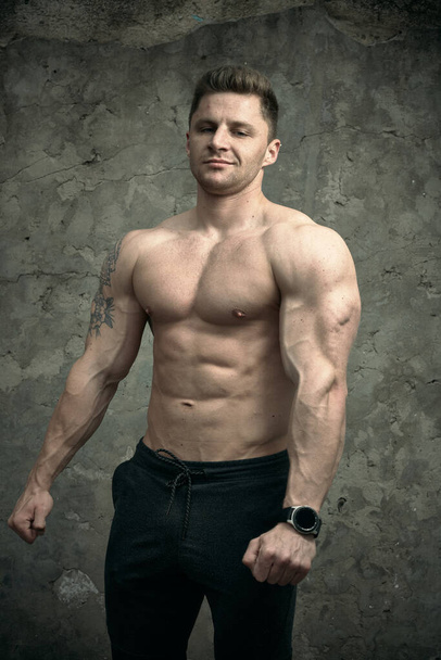 Красивый мускулистый мужчина позирует на фоне бетонной стены. Понятие фитнеса, бодибилдинга, здоровья, образа жизни. Портрет фитнес-модели
. - Фото, изображение