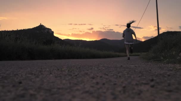 Fiatal lány fut az úton a naplemente alatt a hegyek között. Meleg színek - Felvétel, videó